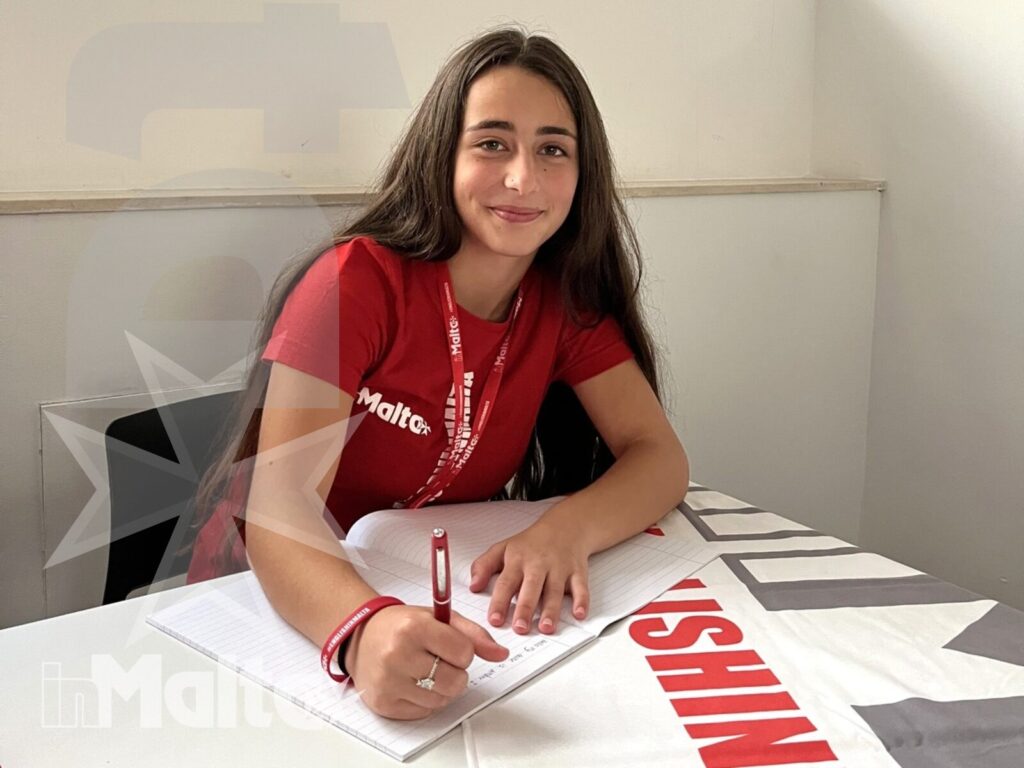 Studentessa sorridente compila un modulo a un tavolo informativo, durante l'accoglienza di un gruppo per un soggiorno linguistico.