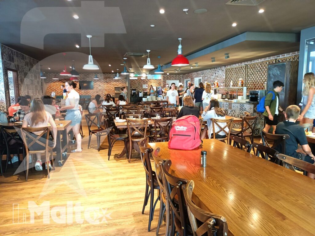 Sala ristorazione affollata con studenti ai tavoli, persone al bancone e lampade colorate, ambiente quotidiano di socialità durante un soggiorno linguistico.
