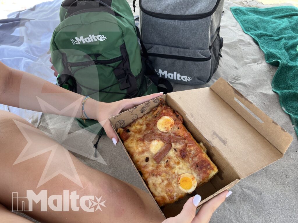 Pizza rettangolare con uova in una confezione aperta, mostrata dall'alto durante una pausa sulla spiaggia tra zaini e teli.