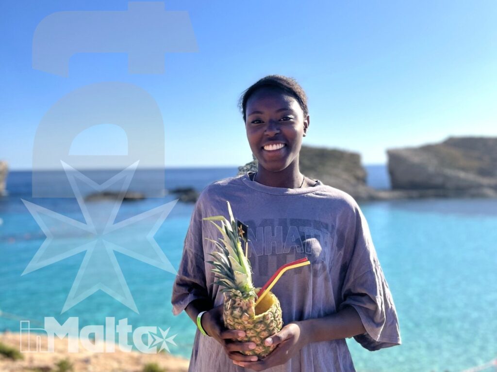 Studentessa inMalta con bevanda sulla baia Studentessa sorridente tiene una bevanda servita in un ananas davanti alla baia turchese durante l'escursione a Malta.