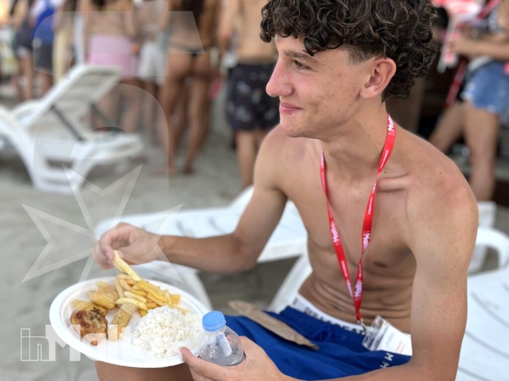 Pranzo degli studenti sulla spiaggia di Mellieha Bay Studente con badge mangia seduto su un lettino, mentre altri giovani condividono il pranzo sulla spiaggia di Mellieha Bay.