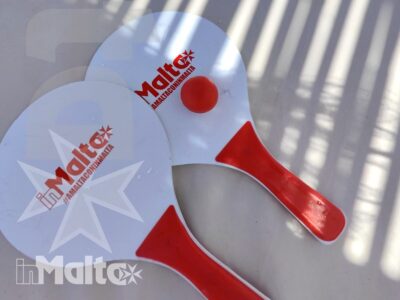 Due palette da ping pong rosse e bianche con logo Malta, illuminate da ombre geometriche sulla superficie.