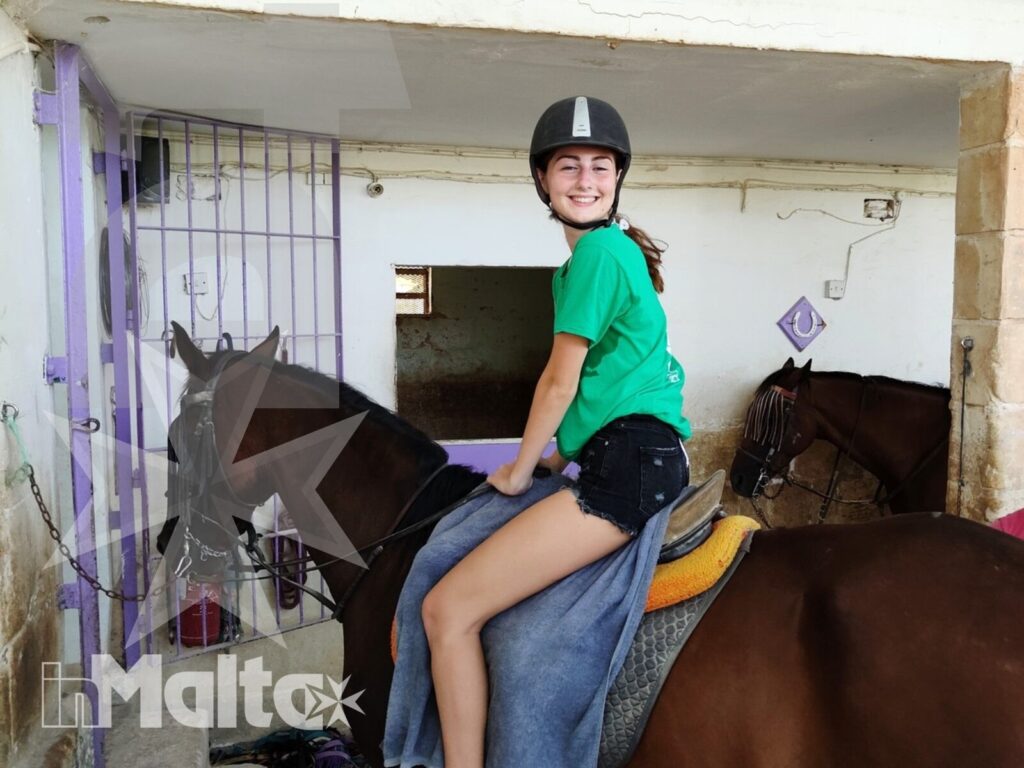 Studentessa sorridente a cavallo, con casco e abbigliamento da equitazione, ritratta all'interno della scuderia inMalta.