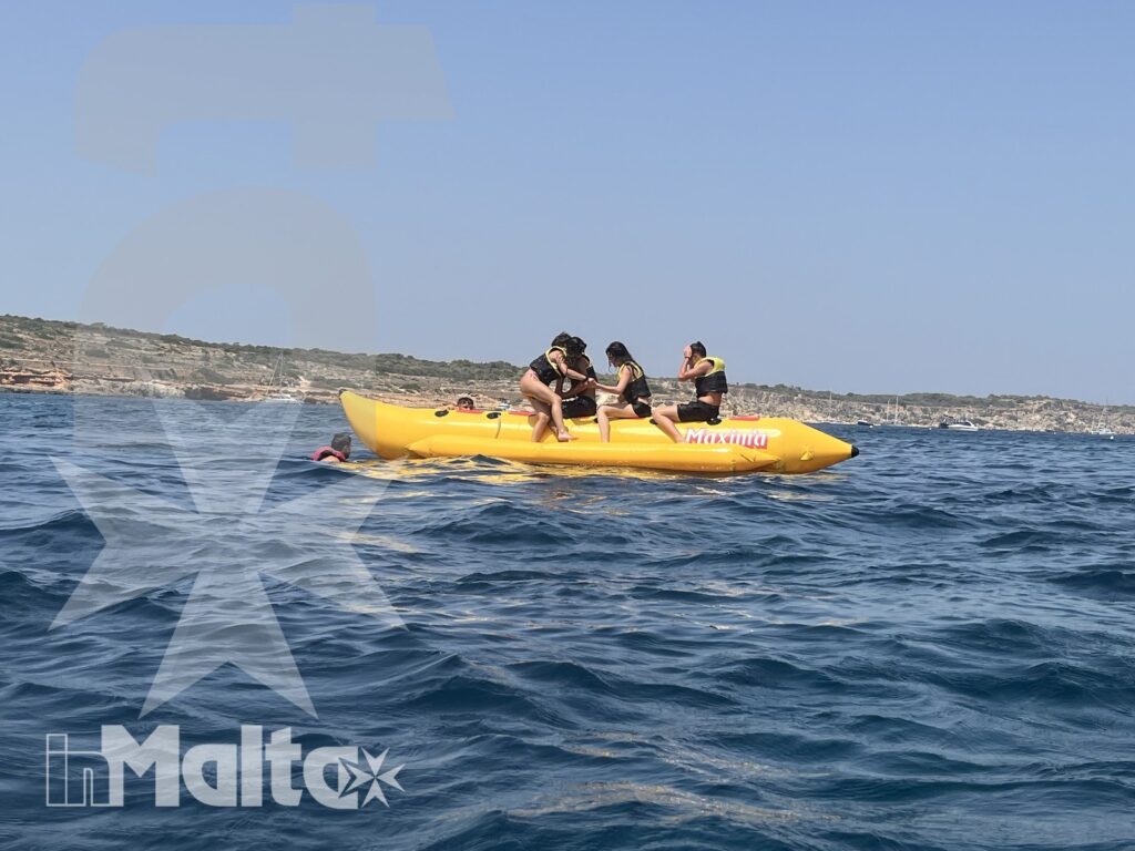 Studenti collaborano su un kayak gonfiabile giallo, pagaiando vicino a una costa rocciosa sotto il cielo sereno.