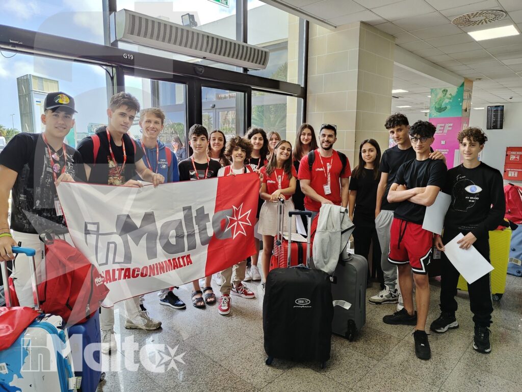 Gruppo internazionale di studenti con valigie e striscione dedicato a Malta, riunito nell'ambiente aeroportuale.