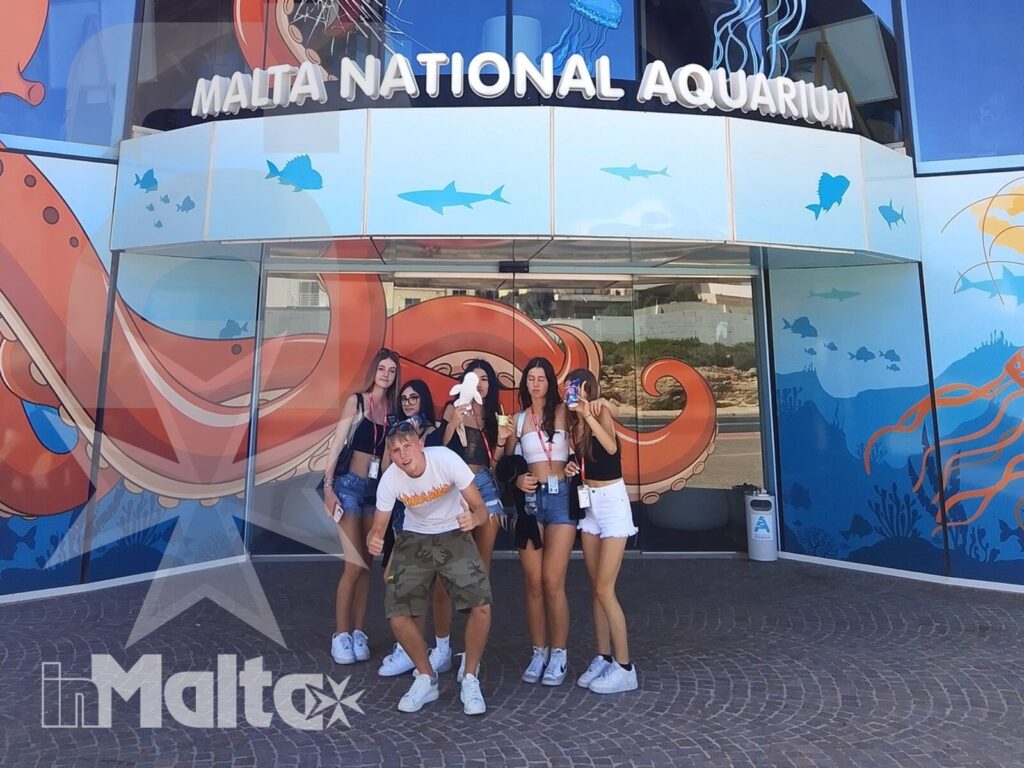 Studenti in visita al Malta National Aquarium durante un soggiorno studio Gruppo di studenti posa davanti all'ingresso decorato del Malta National Aquarium, tra murales marini e facciate vetrate.
