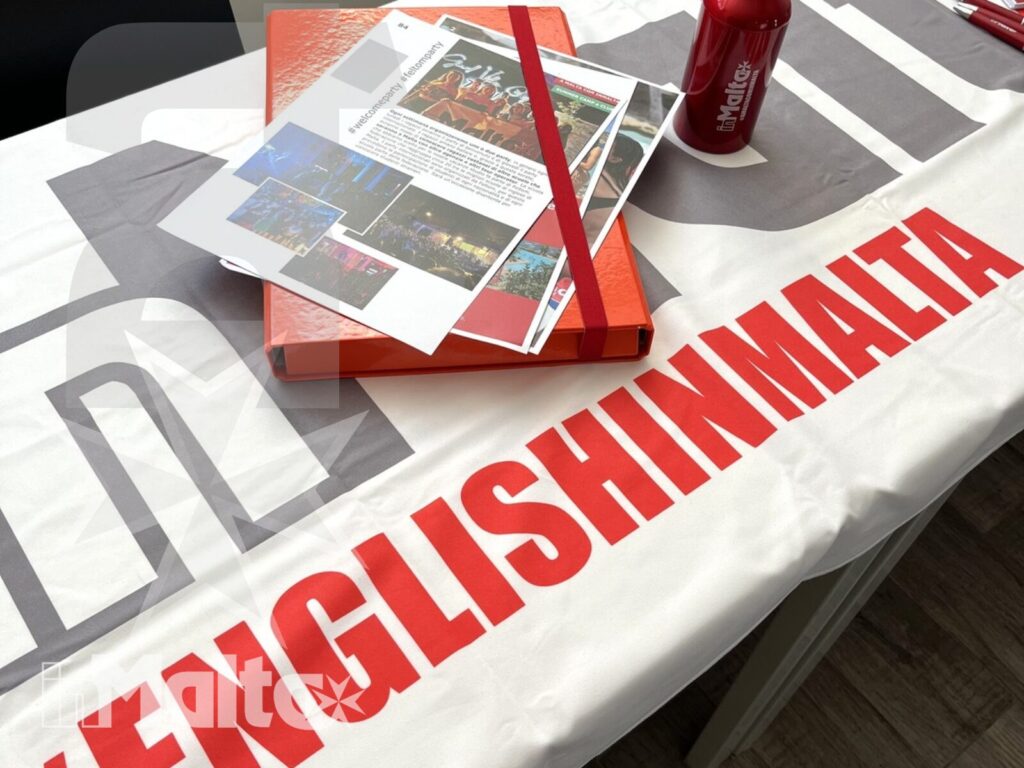 Brochure, quaderni rossi e borraccia sono disposti su un tavolo brandizzato per i partecipanti a un corso d'inglese.