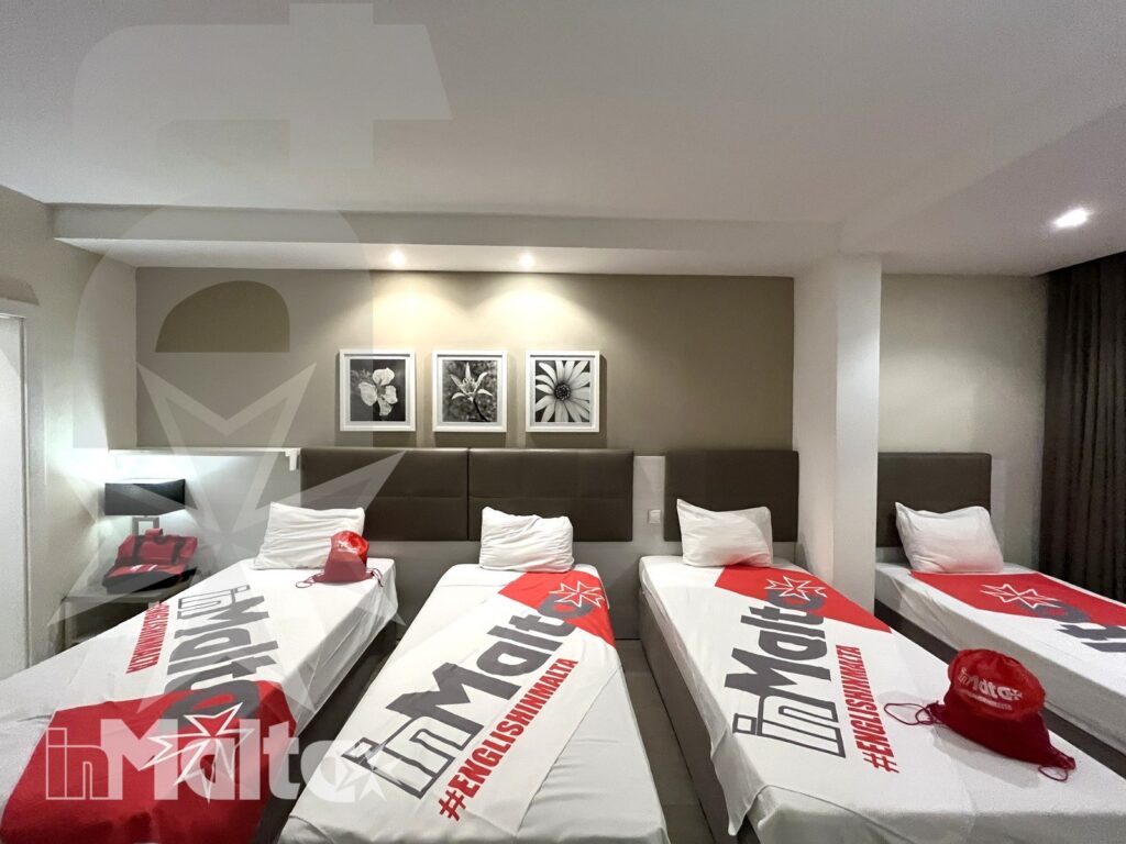 Quattro letti singoli con biancheria coordinata, cappellini e zaini preparano una camera condivisa per studenti in soggiorno studio a Malta.