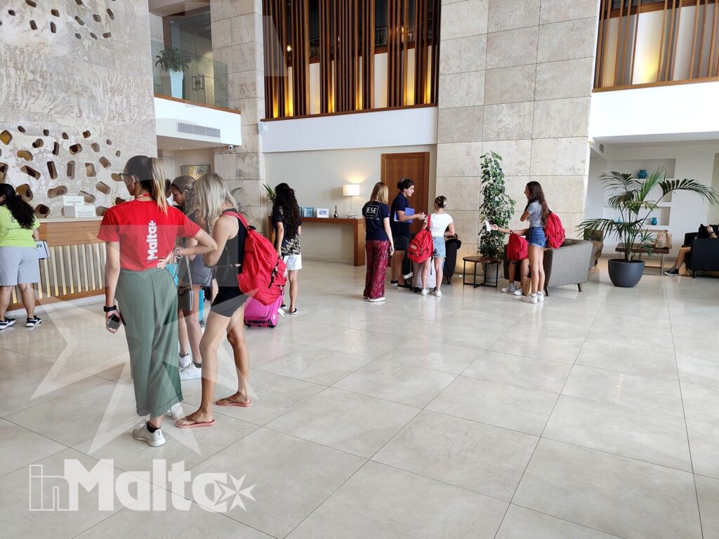 Studenti con zaini e valigie si riuniscono nella hall moderna del resort, circondati da pareti in pietra e arredi luminosi.