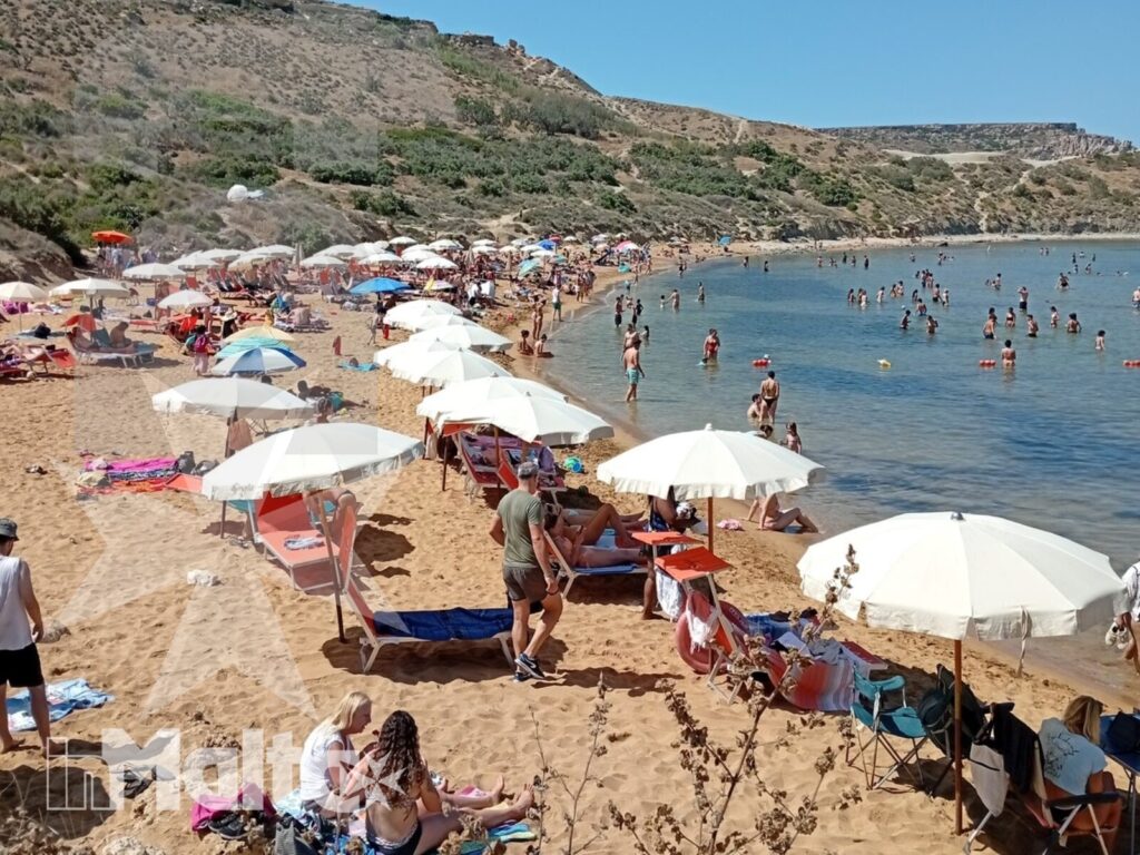 Partecipanti inMalta sulla spiaggia affollata della baia La vista dall'alto mostra una baia maltese affollata, con ombrelloni colorati, bagnanti in acqua e scogliera brulla.