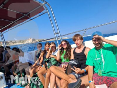 Gruppo di studenti e accompagnatore siede su una barca turistica, circondato dal mare e dalla costa maltese durante l'escursione.