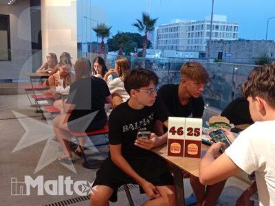 Studenti giocano a carte ai tavoli esterni durante una serata, con vista urbana maltese e atmosfera di socializzazione.
