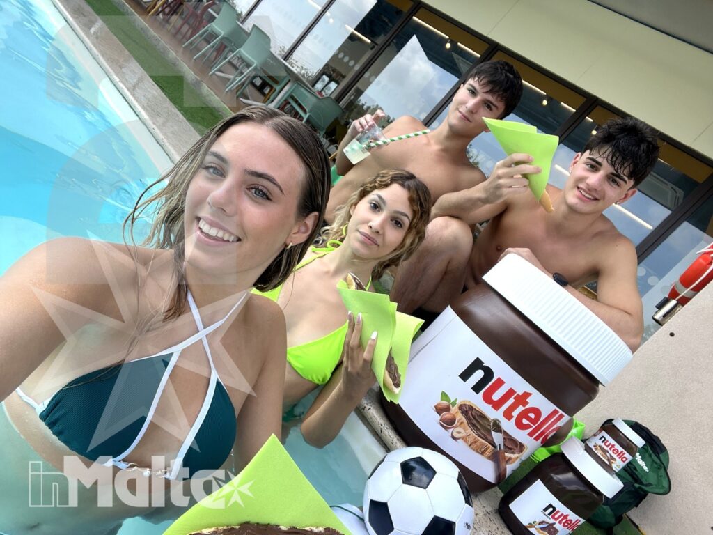 Quattro studenti sorridenti si fotografano in piscina con snack, pallone e crema alla nocciola durante una pausa di gruppo.
