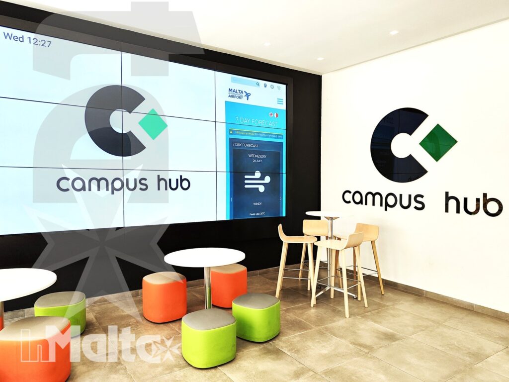 Area comune moderna del Campus Hub con parete digitale, logo, tavoli e sedute colorate per studio e socializzazione.