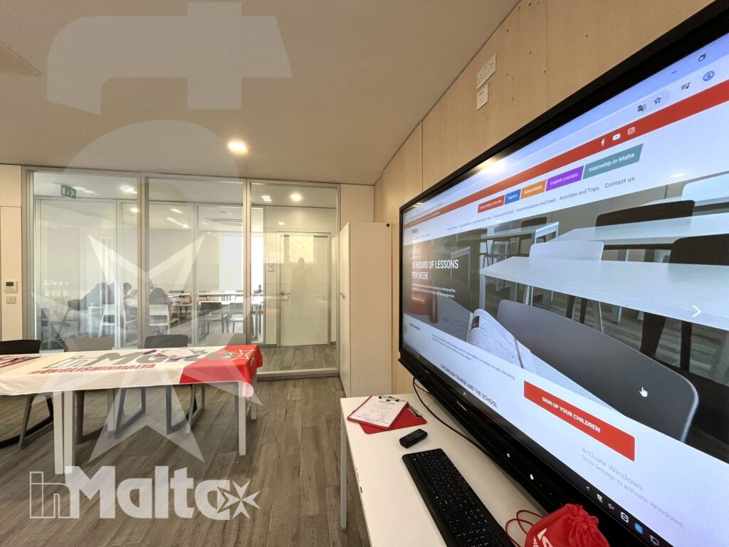Aula moderna con tavoli predisposti, computer e grande schermo digitale, pronta per una sessione di studio dei partecipanti.