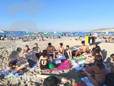 Studenti seduti in cerchio sulla spiaggia svolgono un'attività didattica con quaderni e materiali, tra mare e bagnanti sullo sfondo.