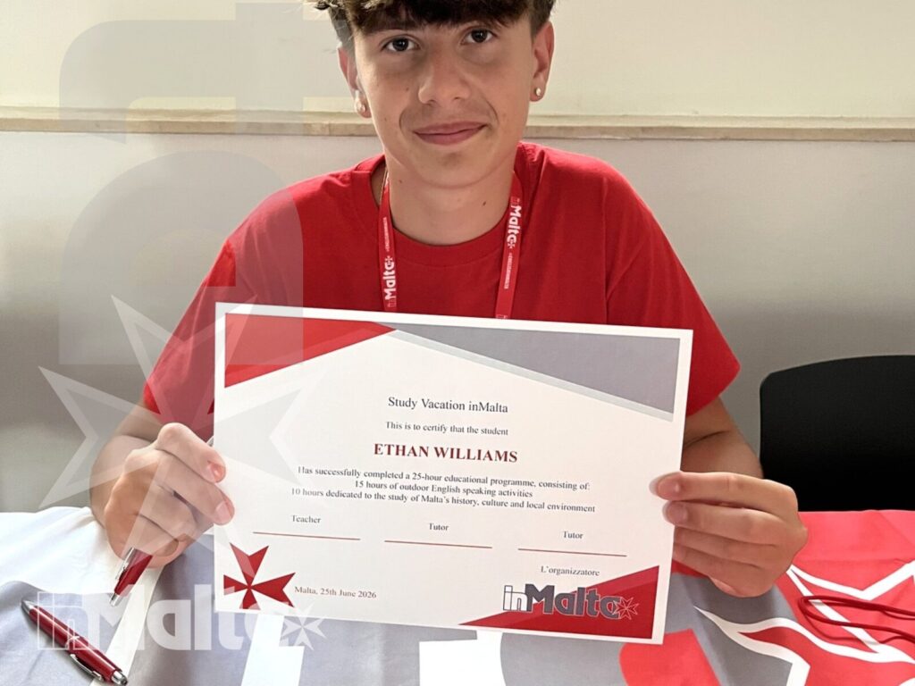 Giovane studente sorridente mostra un certificato seduto a un tavolo brandizzato, celebrando la conclusione del soggiorno studio a Malta.