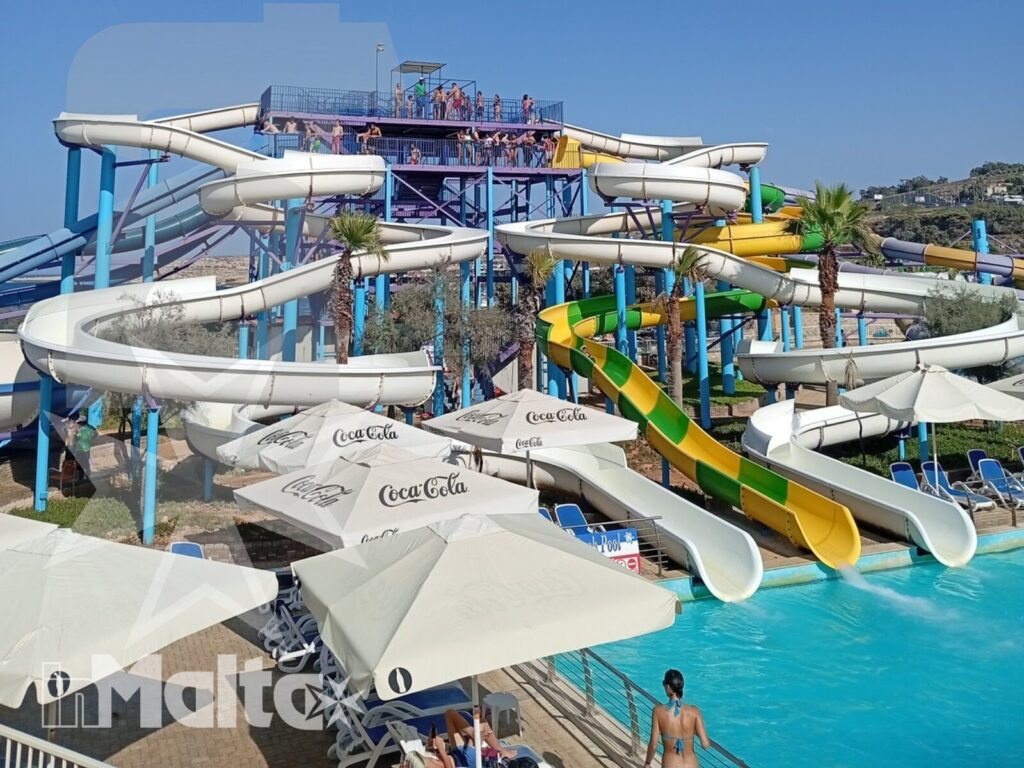 Panoramica di un parco acquatico con scivoli colorati, piscine, ombrelloni e visitatori immersi nell'atmosfera estiva.
