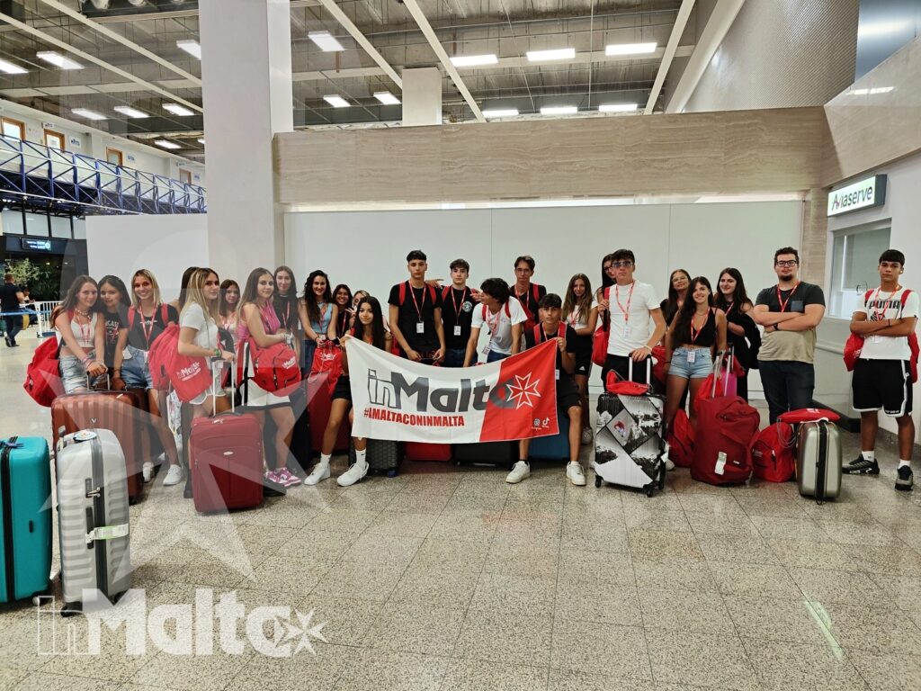 Numerosi studenti con bagagli e accessori rossi posano in aeroporto accanto a uno striscione dedicato a Malta.