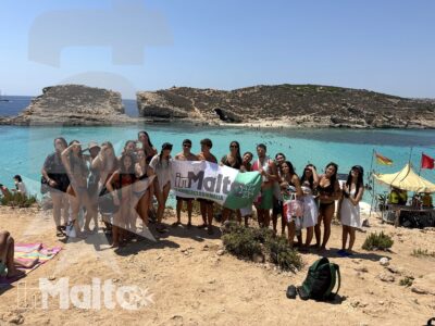 Gruppo internazionale di studenti posa con uno striscione davanti a una baia turchese e alle scogliere calcaree di Malta.