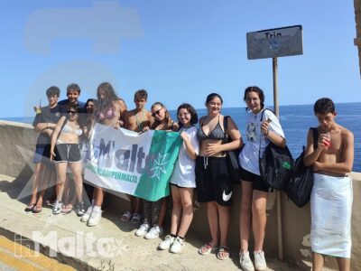 Studenti sorridenti posano con lo striscione del viaggio sul lungomare maltese, immersi nella luce estiva di un'escursione.