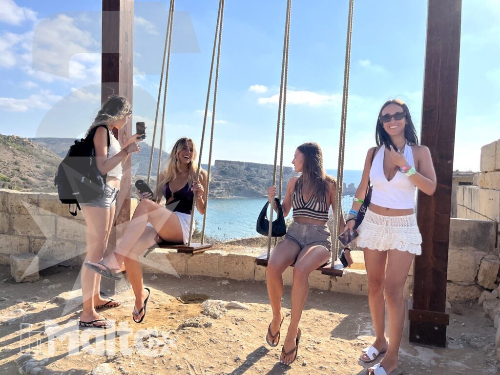 Quattro giovani donne si ritrovano presso le altalene panoramiche sul mare, mentre una partecipante scatta fotografie con lo smartphone.