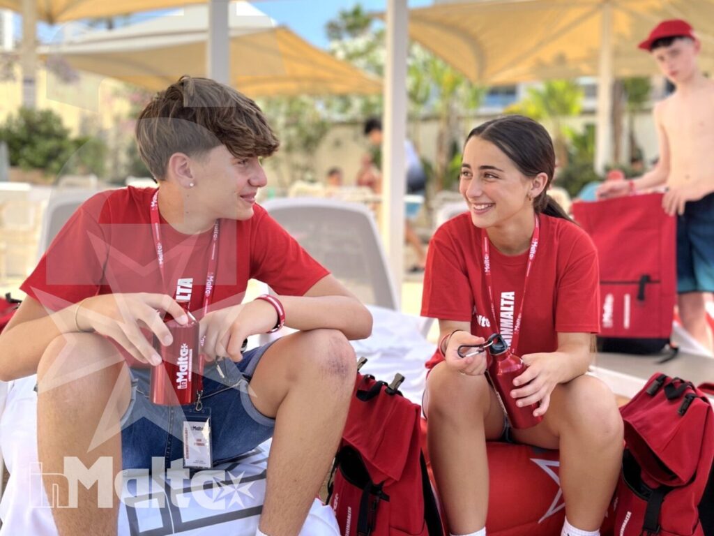 Studenti inMalta si rilassano a bordo piscina Due studenti sorridenti si rilassano a bordo piscina con borse rosse e altri giovani sullo sfondo durante il soggiorno studio.