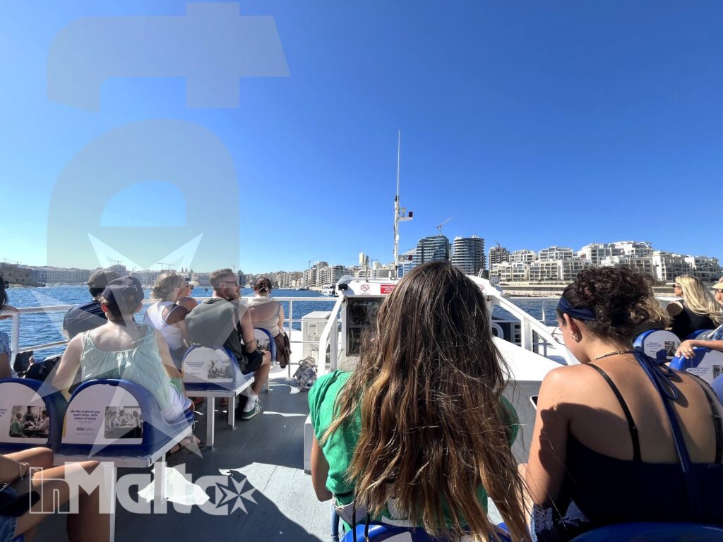 Studenti seduti sul ponte di una barca in navigazione lungo la costa urbana di Malta durante un'escursione sul mare.