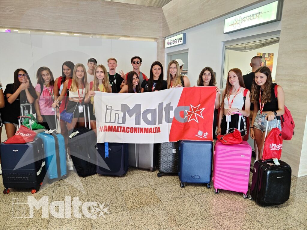 Gruppo internazionale di studenti posa in aeroporto con valigie colorate e striscione dedicato all'accoglienza a Malta.