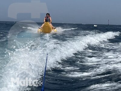 Una persona viene trainata su un gonfiabile giallo, ripresa dalla barca attraverso spruzzi e una lunga scia d'acqua in mare aperto.