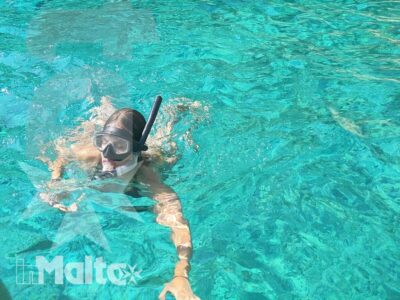 Persona con maschera e boccaglio fa snorkeling davanti a una grotta marina maltese, con altri nuotatori sullo sfondo.