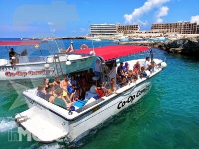 Barca turistica affollata di studenti naviga tra acque turchesi, grotte e costa rocciosa durante un'escursione maltese.