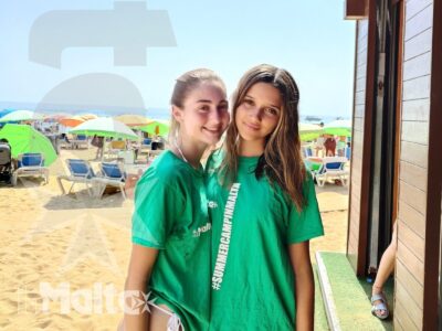 Due studentesse in maglietta verde sorridono davanti alla spiaggia di Golden Bay, ritratte accanto a uno stabilimento balneare.