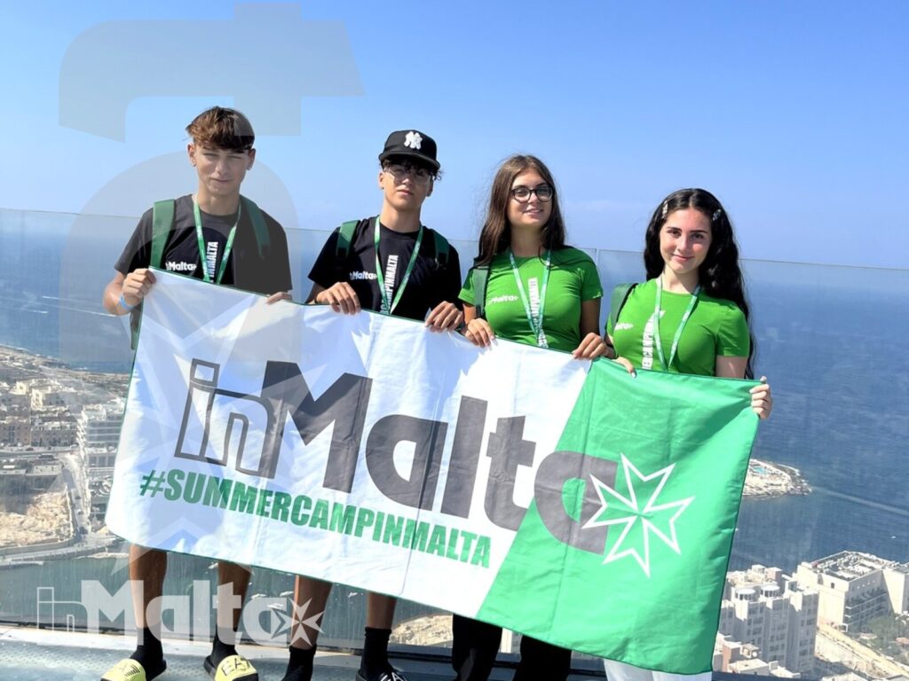 Quattro studenti mostrano un banner del summer camp su una terrazza panoramica affacciata sulla costa di Malta.