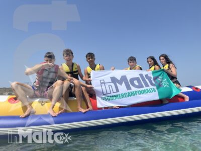 Un gruppo di studenti posa sorridente su un gommone in mare, con giubbotti salvagente e bandiera promozionale dell'attività nautica.