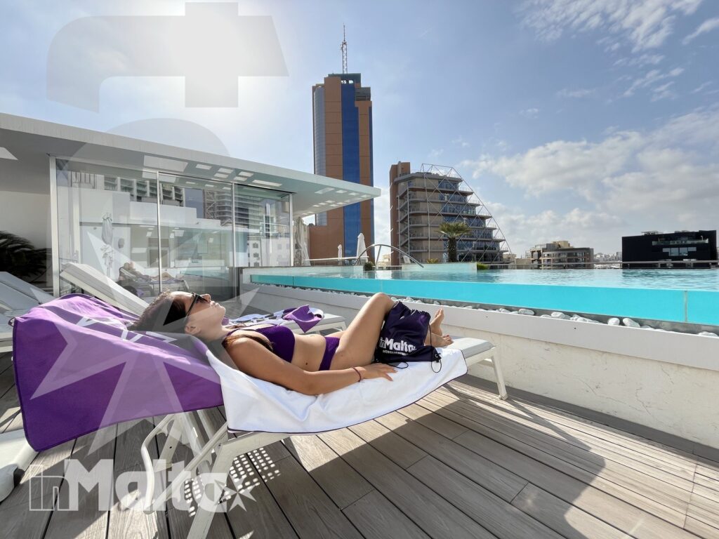 Studente inMalta sul rooftop con piscina panoramica Studente rilassato su un lettino accanto alla piscina panoramica sul tetto, con vista urbana e terrazza soleggiata a Malta.