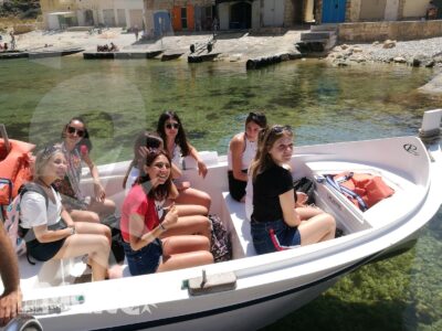 Studenti seduti su una barca condividono una gita nelle acque limpide di Malta, con la rada costiera visibile alle spalle.