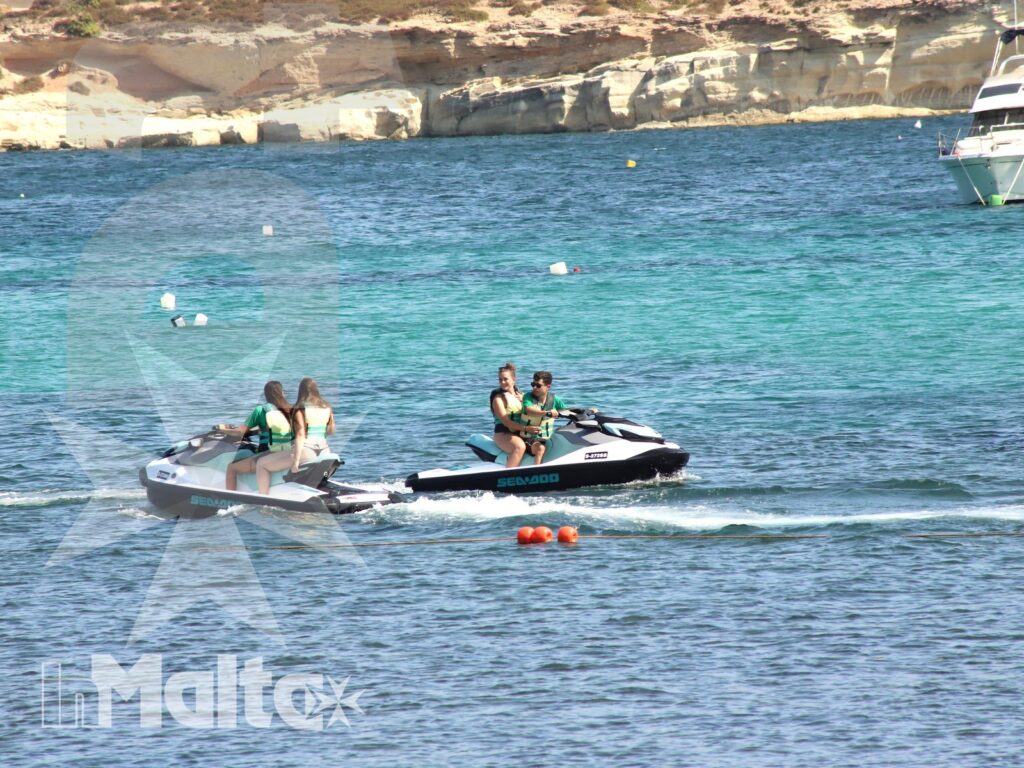 Due moto d'acqua attraversano una baia turchese con scie parallele, scogliere chiare e barche ormeggiate sullo sfondo.