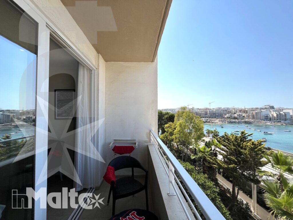 Balcone arredato con sedie e tavolino, affacciato su una baia mediterranea, edifici costieri e vegetazione del paesaggio maltese.