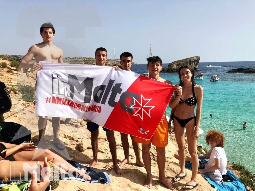Studenti inMalta sulla costa rocciosa con bandiera maltese Gruppo di studenti posa con una bandiera maltese sulla spiaggia rocciosa, con il mare turchese sullo sfondo.