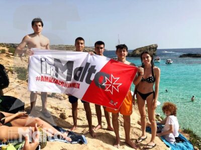 Gruppo internazionale di studenti posa con una bandiera maltese su una spiaggia rocciosa, tra mare turchese e atmosfera conviviale.