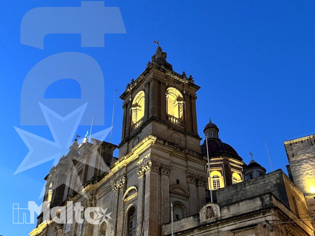 Chiesa barocca illuminata nelle città storiche maltesi Chiesa barocca monumentale con torre campanaria illuminata, osservata dal basso durante una visita serale alle città storiche di Malta.