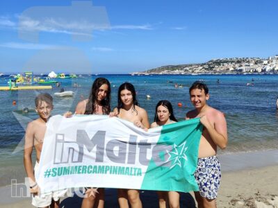 Sei studenti sorridenti mostrano lo striscione del programma sulla spiaggia, con il mare maltese alle loro spalle.
