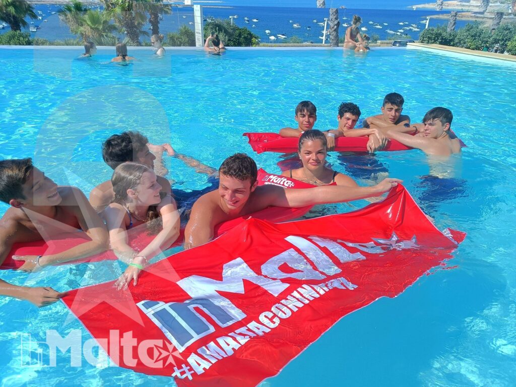 Gruppo di studenti partecipa all'attività in piscina Gruppo numeroso di studenti sostiene uno striscione rosso nell'acqua della piscina, con il mare visibile sullo sfondo.
