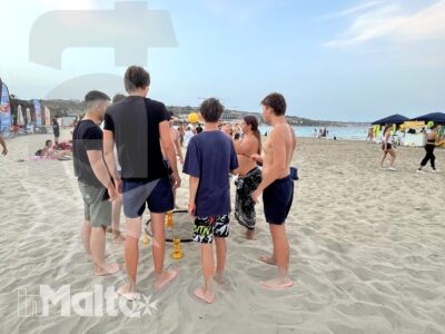 Gruppo di studenti riunito sulla spiaggia, con il mare, gli ombrelloni e altri bagnanti sullo sfondo di una giornata a Malta.