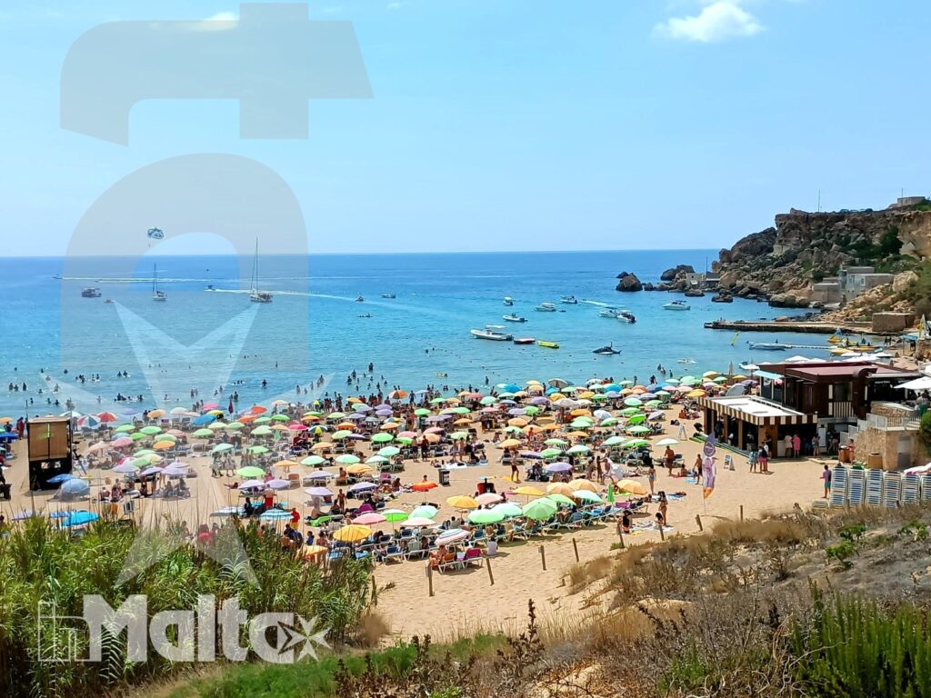 Baia di Golden Bay affollata durante una giornata di mare, con ombrelloni multicolori, bagnanti, imbarcazioni e scogliera rocciosa.