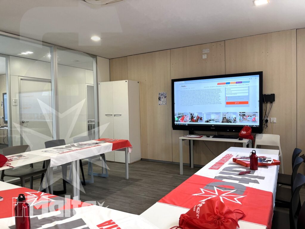 Sala polifunzionale vuota con tavoli a ferro di cavallo, borracce, materiali informativi e schermo digitale, pronta per un'attività collettiva.