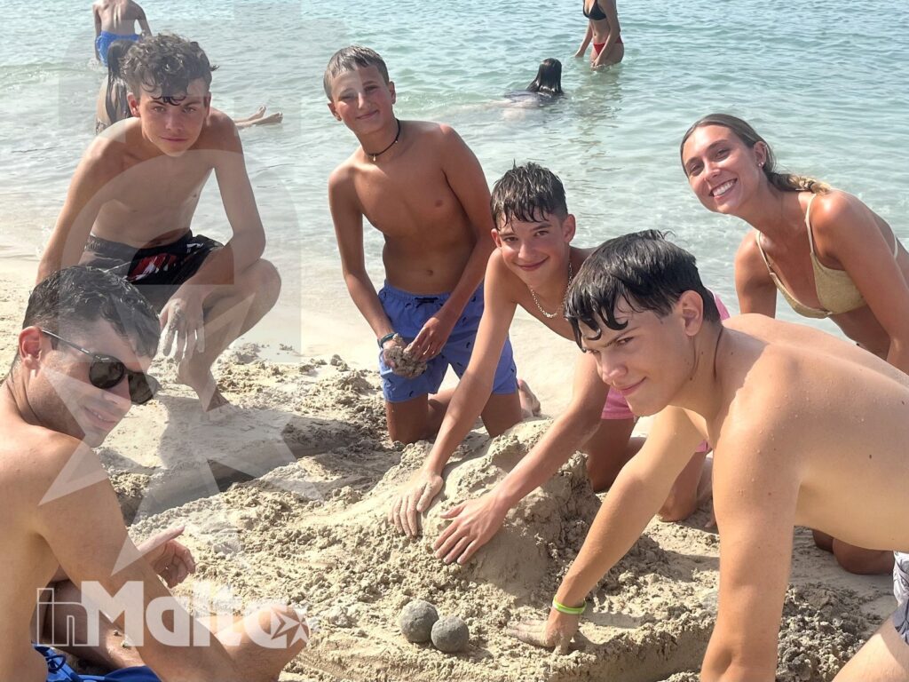 Studenti e accompagnatori partecipano a un'attività creativa sulla sabbia, con il mare limpido di Santa Maria sullo sfondo.