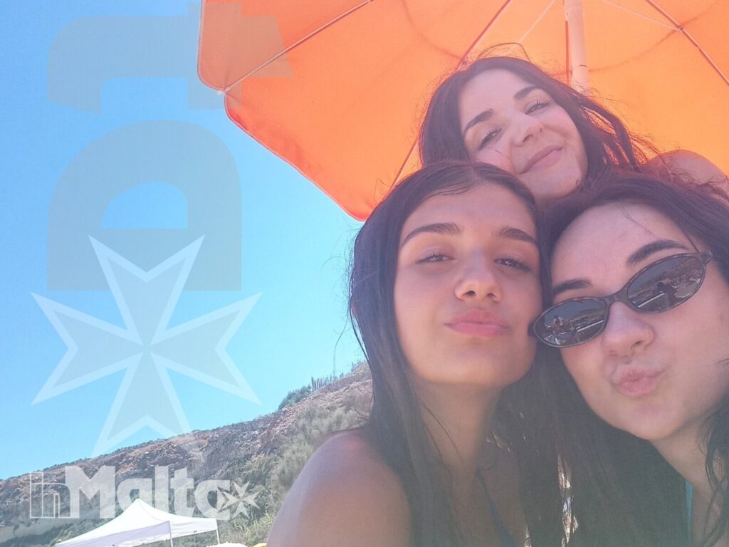 Studentesse inMalta condividono un momento in spiaggia Tre studentesse sorridono sulla spiaggia sotto un ombrellone arancione, con la costa rocciosa sullo sfondo durante una vacanza studio.