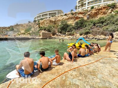 Gruppo di studenti in costume utilizza un pedalò e una piattaforma nelle acque limpide di Golden Bay, davanti alla costa maltese.